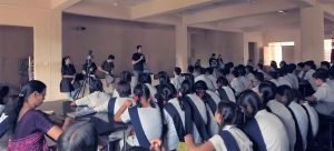 There are many men and women standing in the front. One man is talking. In front of them, many students are sitting. They are wearing their school uniforms.