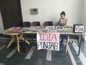 A table stacked with posters and phulkaris. A poster saying IDIA Punjab is pasted on the table. A girl is sitting on the other side of the table.