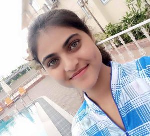 A girl is standing and smiling towards the camera. She is wearing white and blue checked clothes. Behind here, there is a building and a pool.